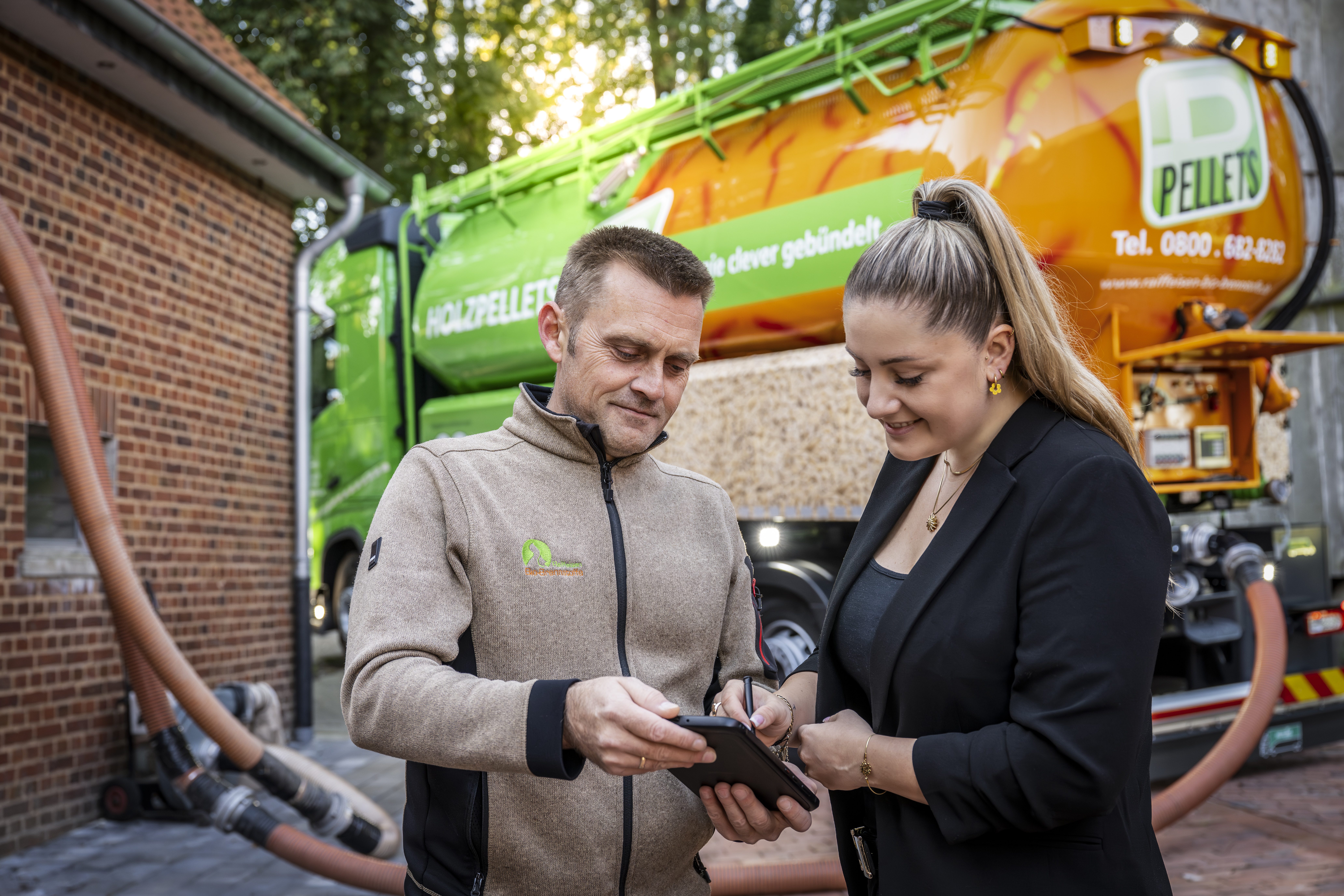 Zwei Personen stehen vor einem Holzpellets‑Lieferfahrzeug und bedienen gemeinsam ein mobiles Endgerät mit Eingabestift.