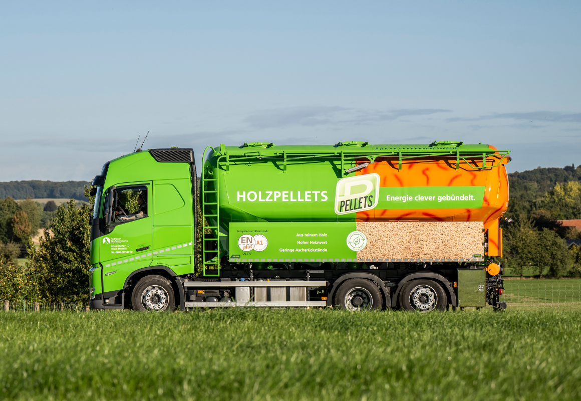 Grüner LKW mit Holzpellet-Aufbau steht auf einer Wiese, ländliche Landschaft im Hintergrund.