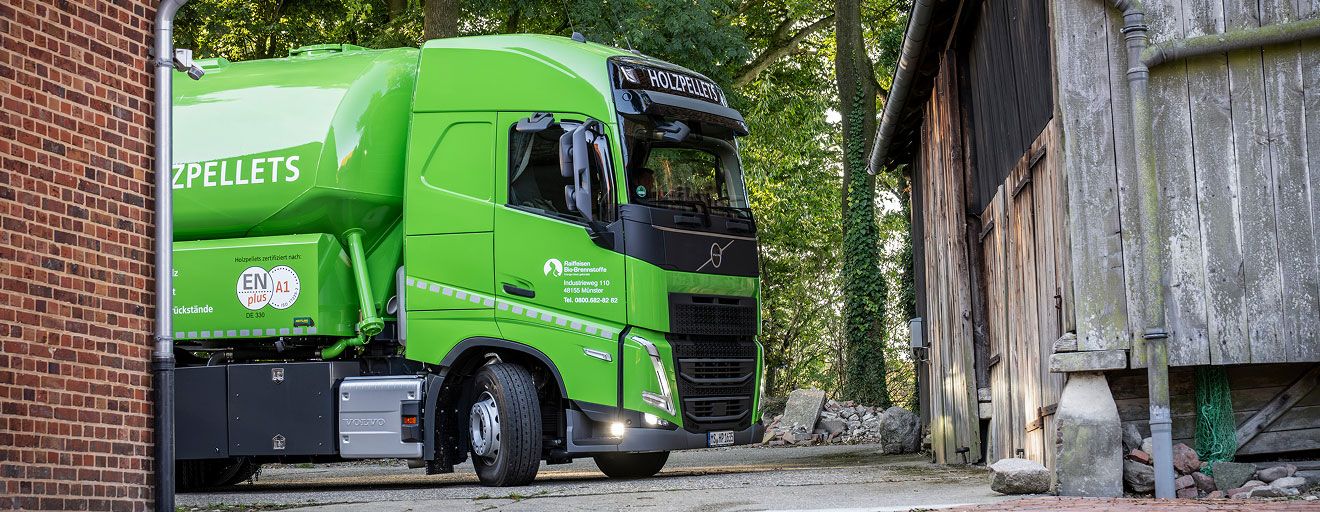 Grünes Holzpellets‑Silofahrzeug fährt zwischen zwei Gebäuden hindurch, umgeben von Bäumen und Hofpflaster.