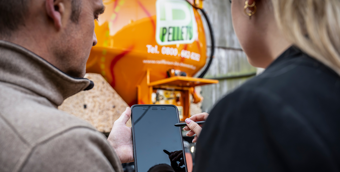 Zwei Personen stehen vor einem Holzpellets‑Lieferfahrzeug und bedienen gemeinsam ein mobiles Endgerät mit Eingabestift.