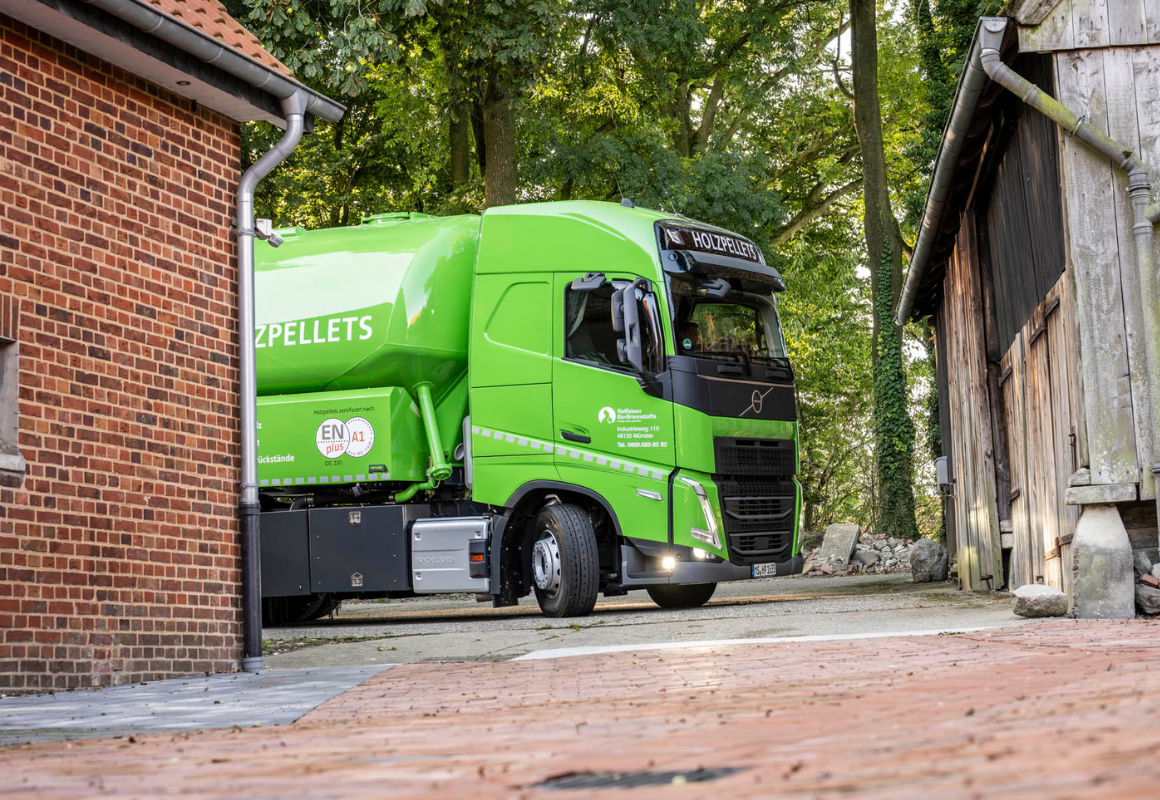 Grüner Silofahrzeug-LKW für Holzpellets fährt zwischen zwei Gebäuden hindurch und erreicht eine Hofeinfahrt.