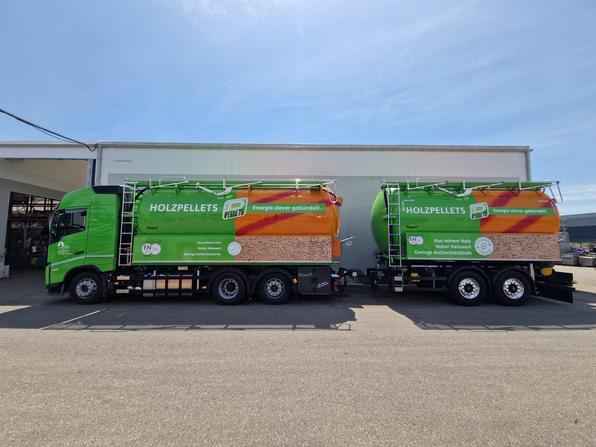 Zwei gekuppelte grüne Holzpellets‑Silofahrzeuge stehen seitlich vor einer Halle, beide mit großflächiger Beschriftung zu Holzpellets und Produktinfos