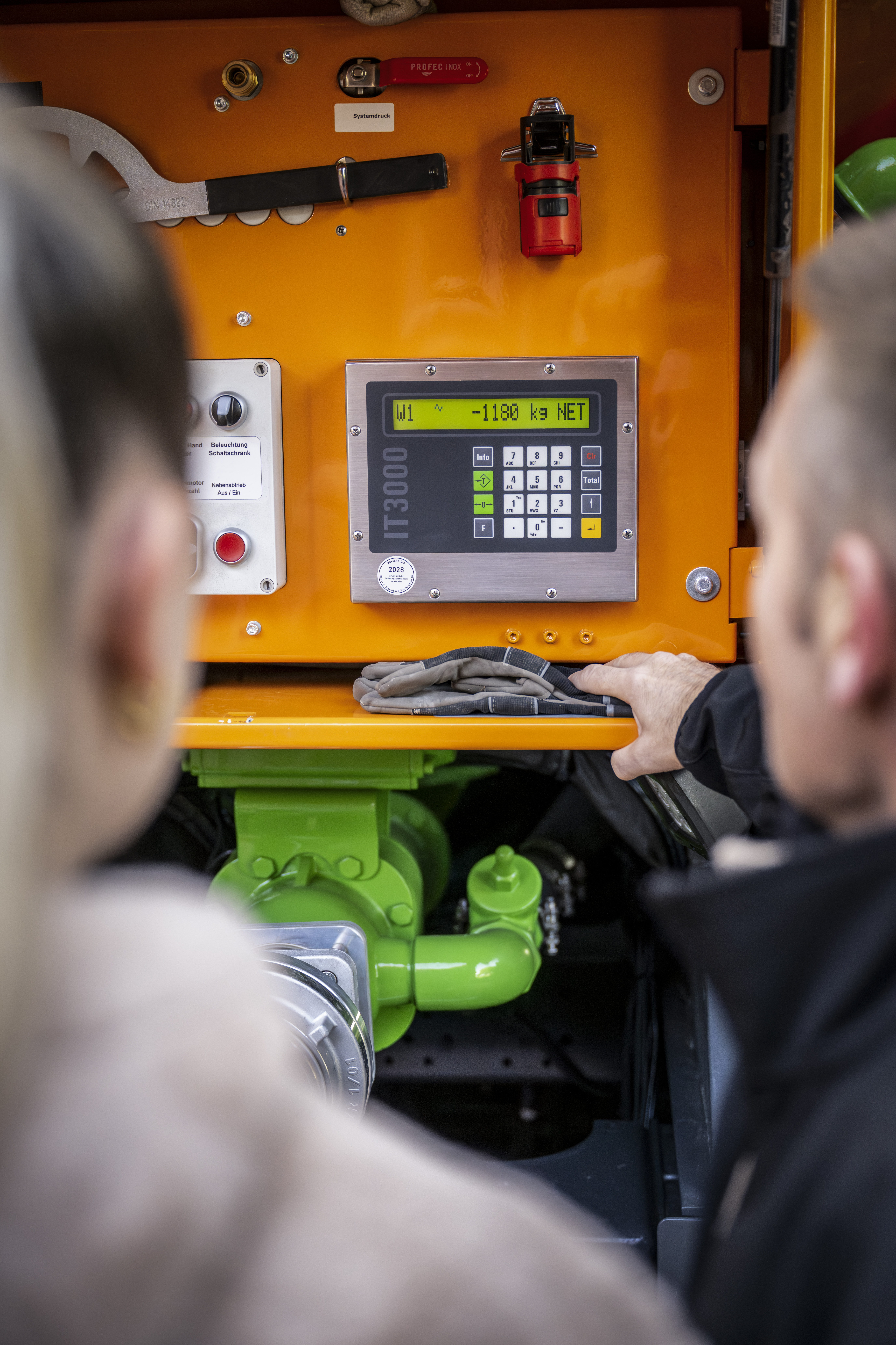 Bedienfeld eines Holzpellets‑Silofahrzeugs mit digitaler Anzeige und Steuerung, zwei Personen stehen davor und betrachten die Technik.