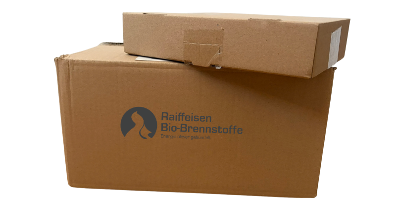 Zwei gestapelte Versandkartons aus brauner Wellpappe, einer davon mit dem Raiffeisen Bio-Brennstoffe Logo.