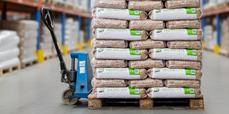 Darstellung einer Palette mit gestapelten Holzpelletsäcken in einer Lagerhalle. Ein Hubwagen steht vor der Palette. Das Bild enthält teilweise KI‑generierte Elemente.