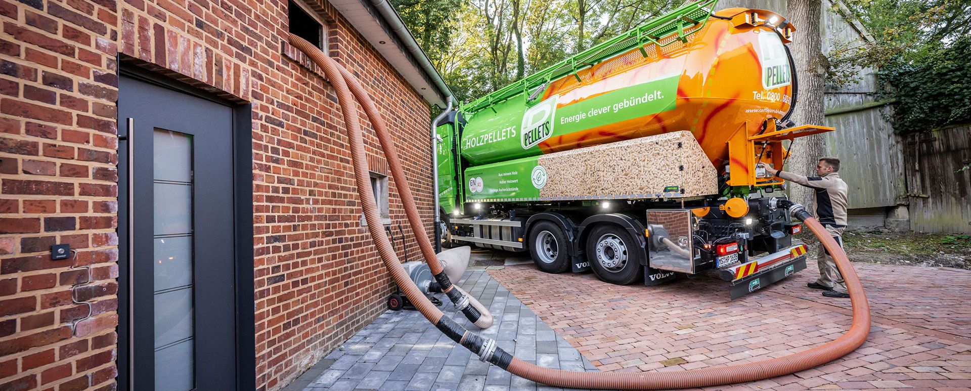 Pellets-Silo-LKW bei der Befüllung eines Hauses, mit angeschlossenen Förderschläuchen und Absauggebläse an der Außenwand.