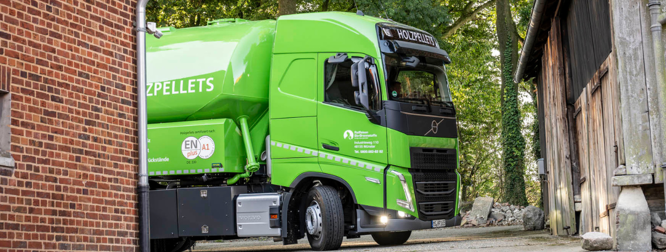 Grüner Holzpellet‑LKW von Raiffeisen Bio‑Brennstoffe fährt auf einen Hof zwischen einem Ziegelgebäude und einer Holzscheune.