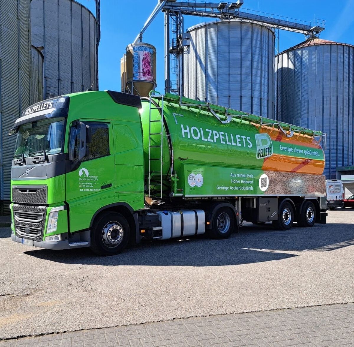 Grüner Silowagen für die Lieferung von Holzpellets, geparkt vor großen Lagersilos auf einem Betriebshof. Auf dem Fahrzeug sind Produktbeschriftungen und Pellet-Motive zu sehen.